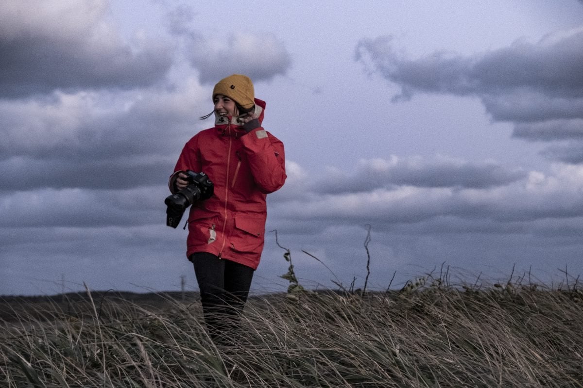 A woman wearing a red jacket and a yellow beanie holding a camera in a field of tall grass, smiling and looking off to the side