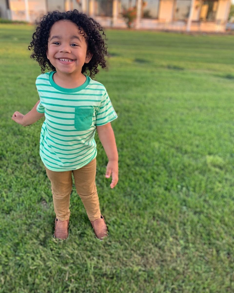 A young boy wearing a green striped shirt and tan pants smiling in a field of grass