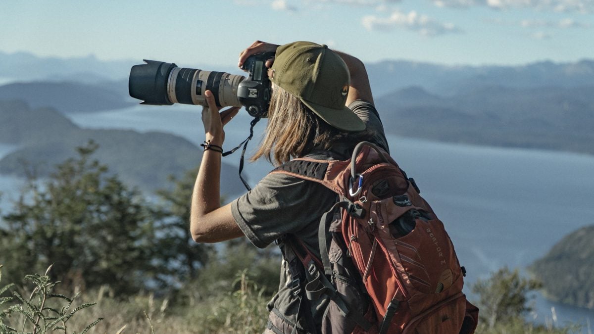 A woman wearing a green hat backwards and a red Osprey pack, holding a camera to take a shot.