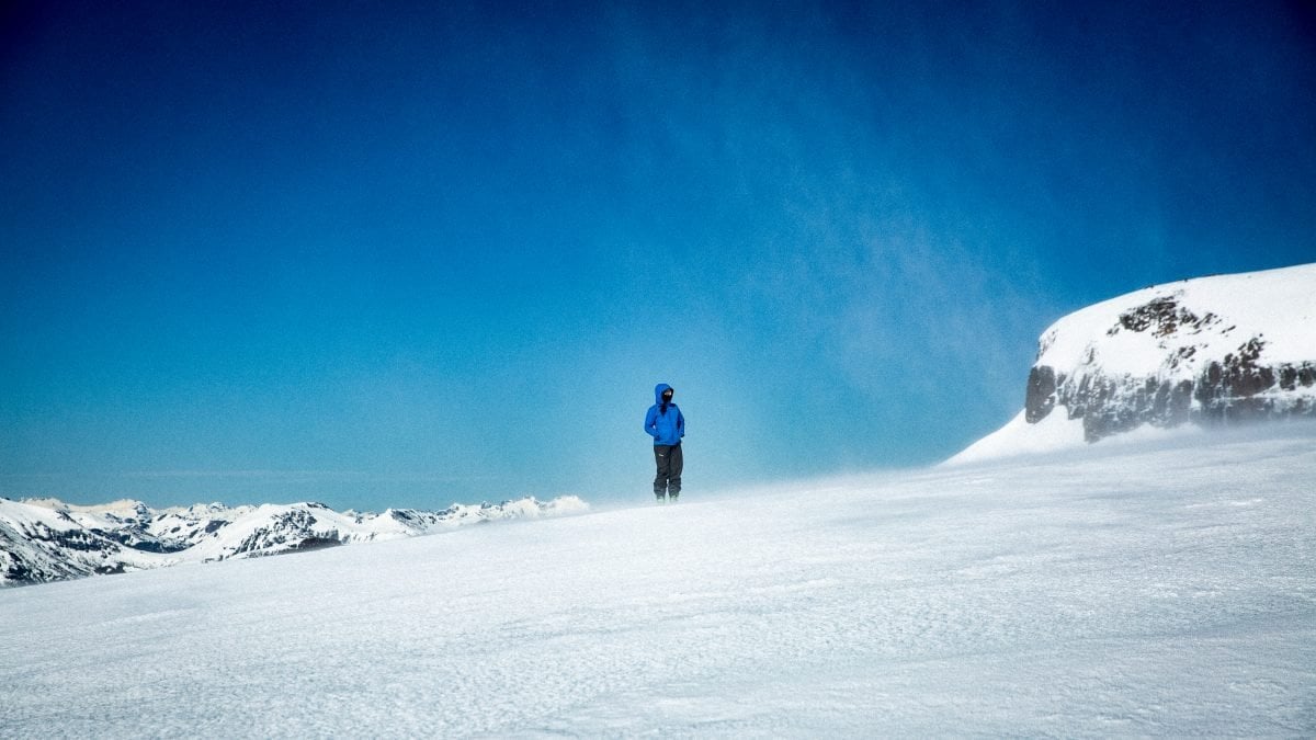 A person wearing a blue jacket with the hood up stands on a snowy mountain with their hands in their pockets