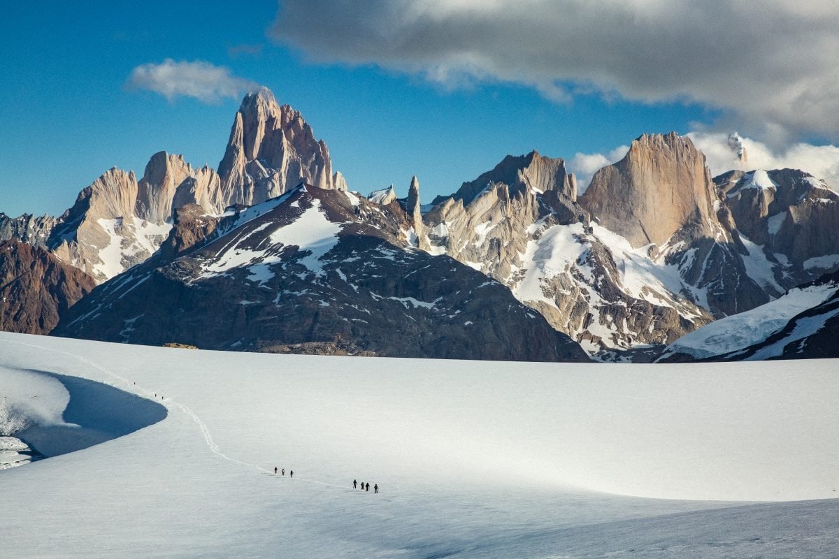 Two small groups of people waling along a snowy trail, with stark, jagged, snowy mountain peaks in the distance