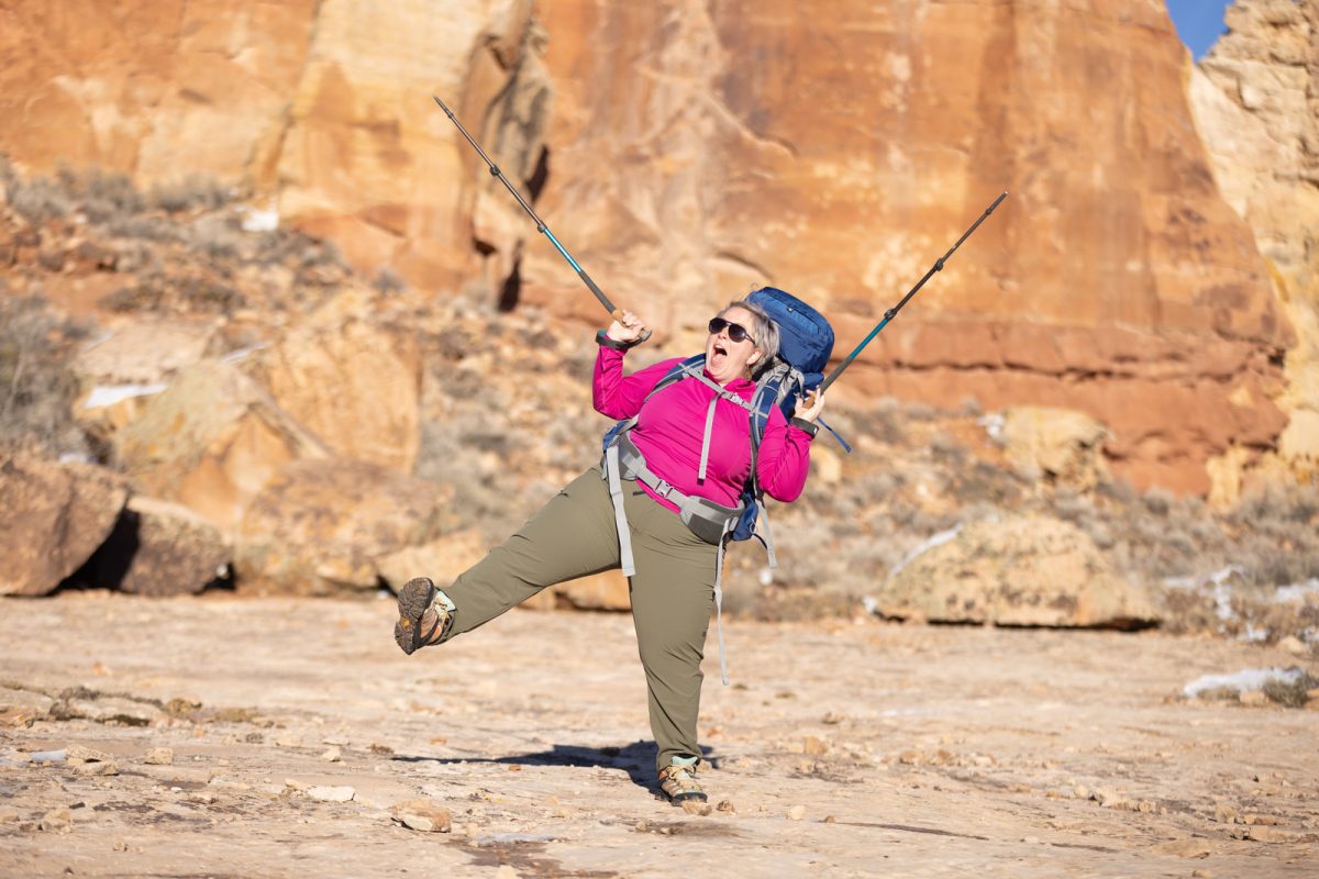 A woman wearing backpacking gear holding her hiking poles up in celebration while lifting one leg off the ground