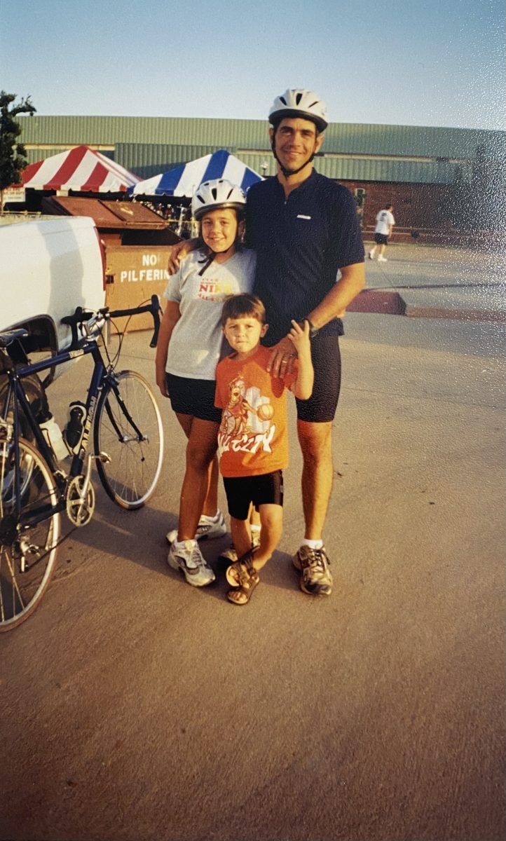 Two young children, one wearing a bike helmet, standing next to an adult wearing a bike helmet