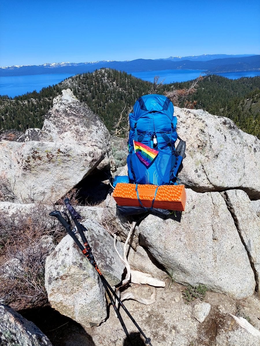 A blue backpack, with an orange sleeping pad and a pride flag strapped to the outside