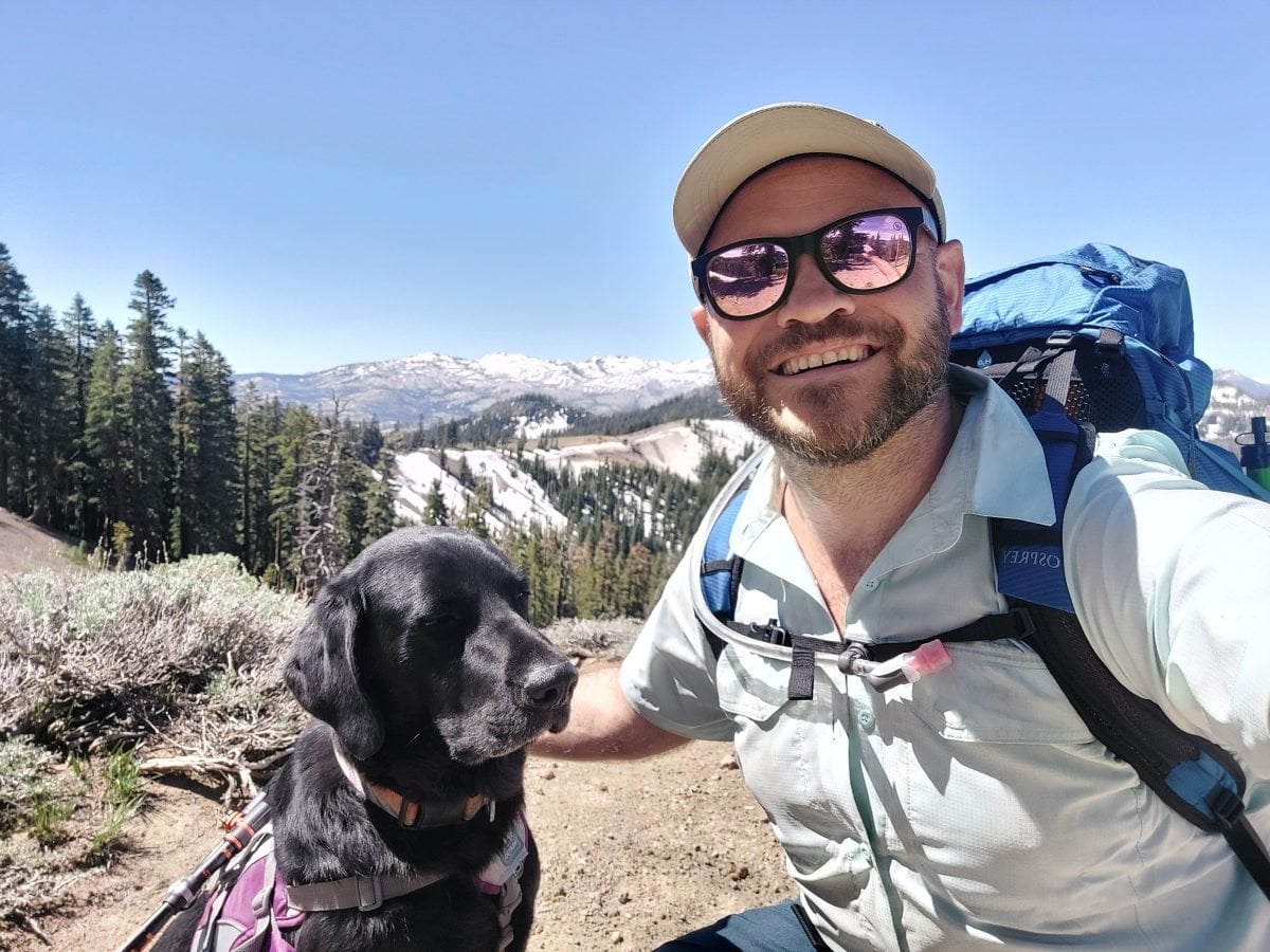 A man in backpacking gear posing with his black lab along the Tahoe Rim Trail