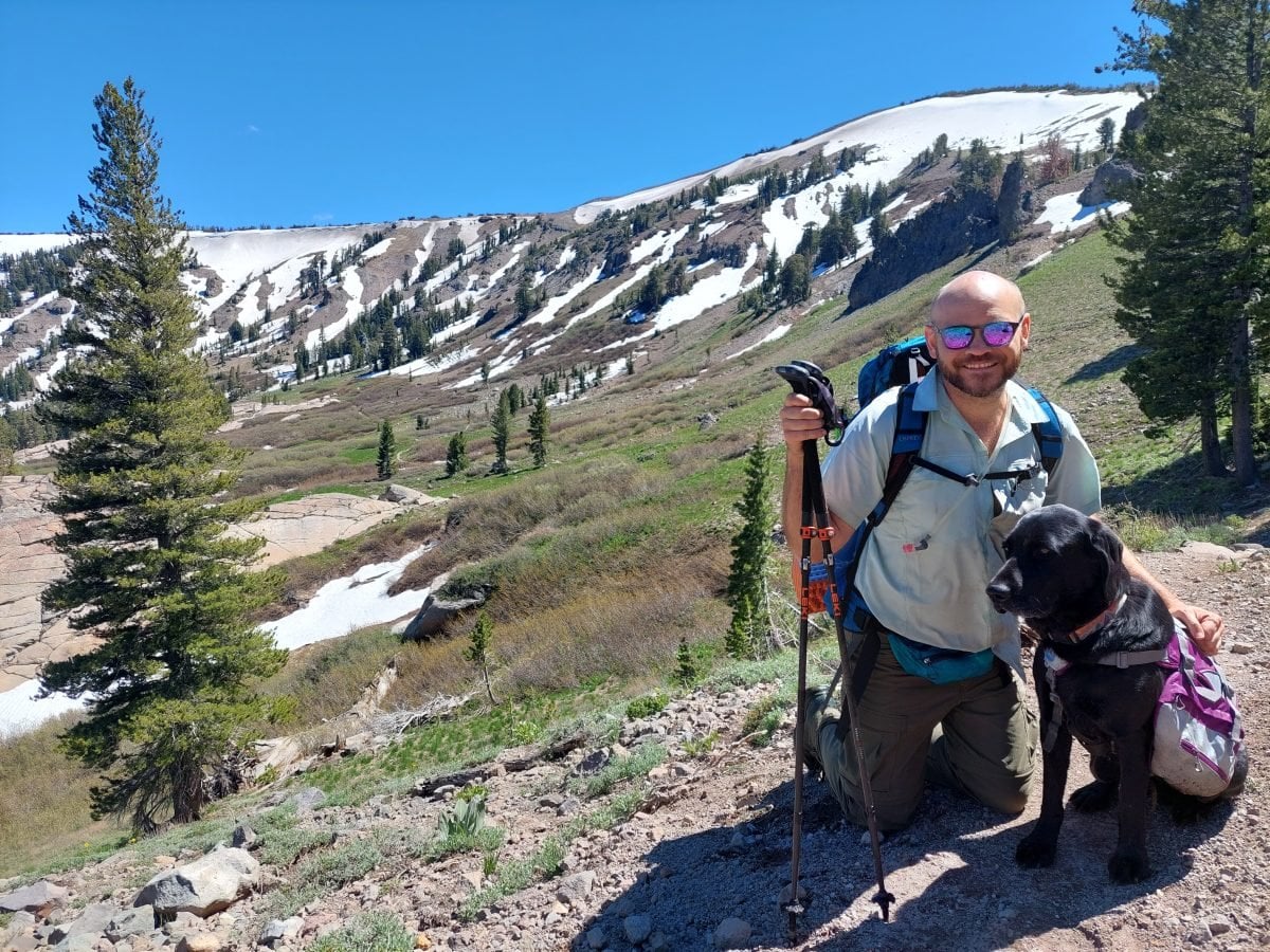 A man in backpacking gear kneeling next to his black lab on the Tahoe Rim Trail