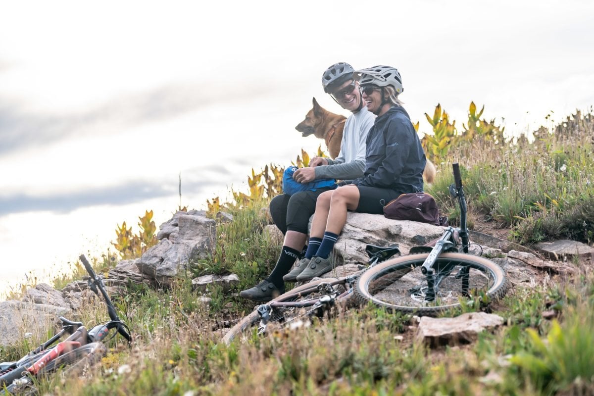 A man, woman and dog sitting on a rock, looking out in the distance, with bikes laying beside them