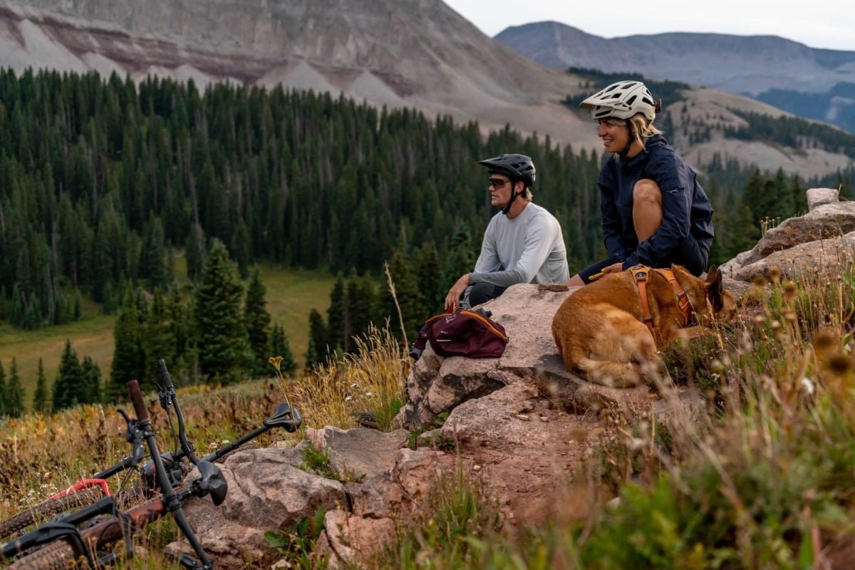 A man, woman and dog sitting on rocks aside a trail, mountain bikes laying beside them
