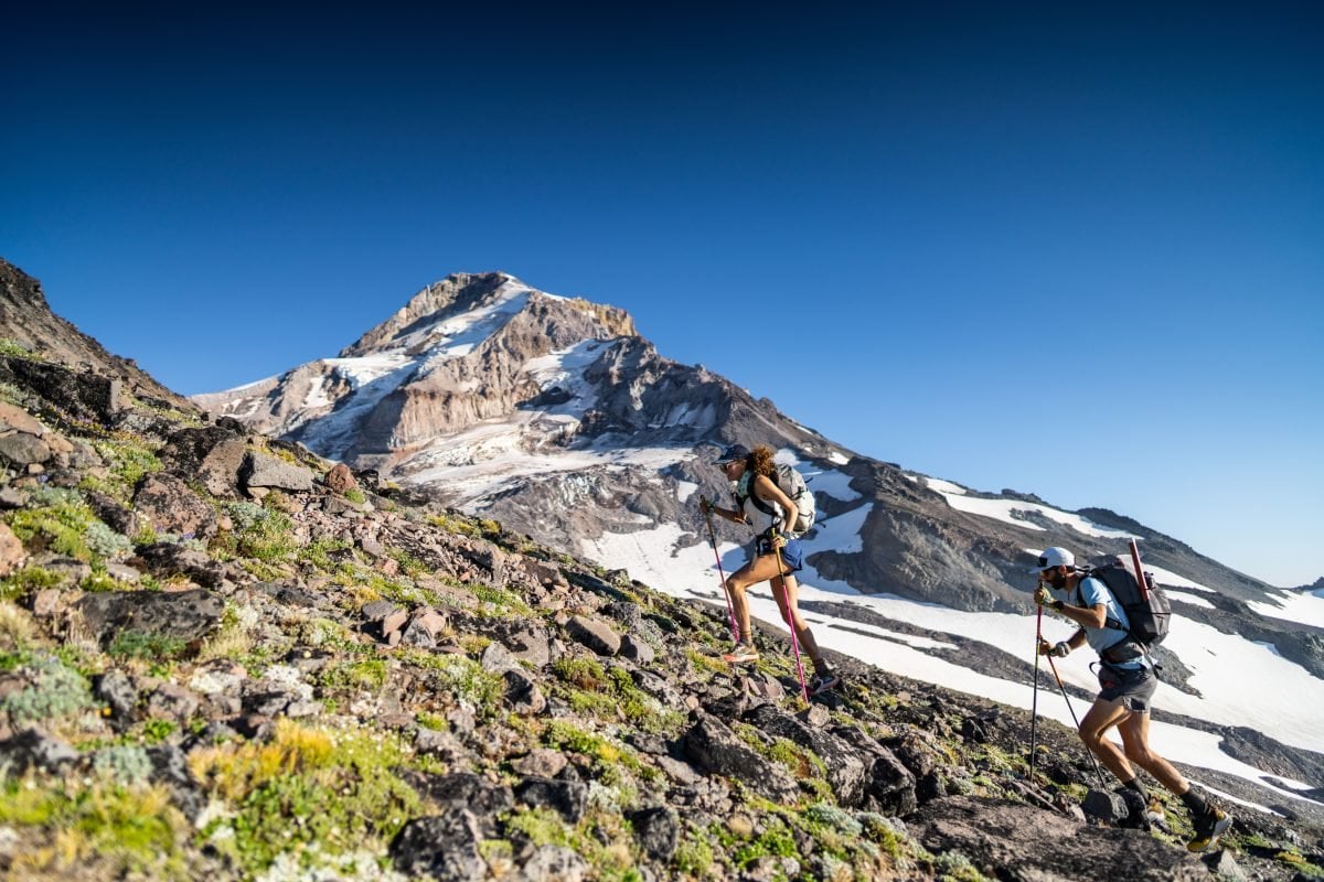 A man and woman trail running up a mountain, with snowy peaks in the backgroun