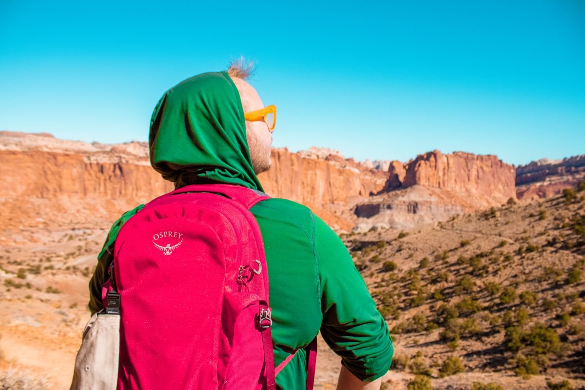 A man wearing a green sweatshirt, red Osprey backpack and yellow glasses, looking out over a desert landscape.