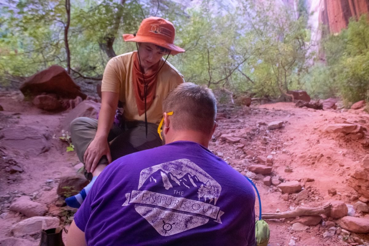 Two people sitting down along a desert trail, looking at something on a camera, with the person facing away from the camera wearing a purple TVOP shirt