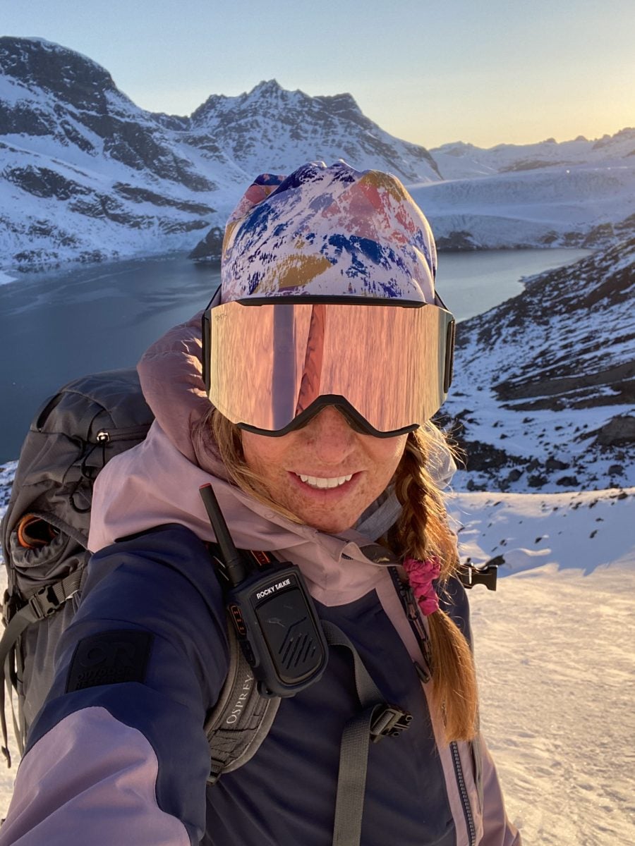 A woman standing on a snowy peak wearing ski gear, with water and mountains in the background
