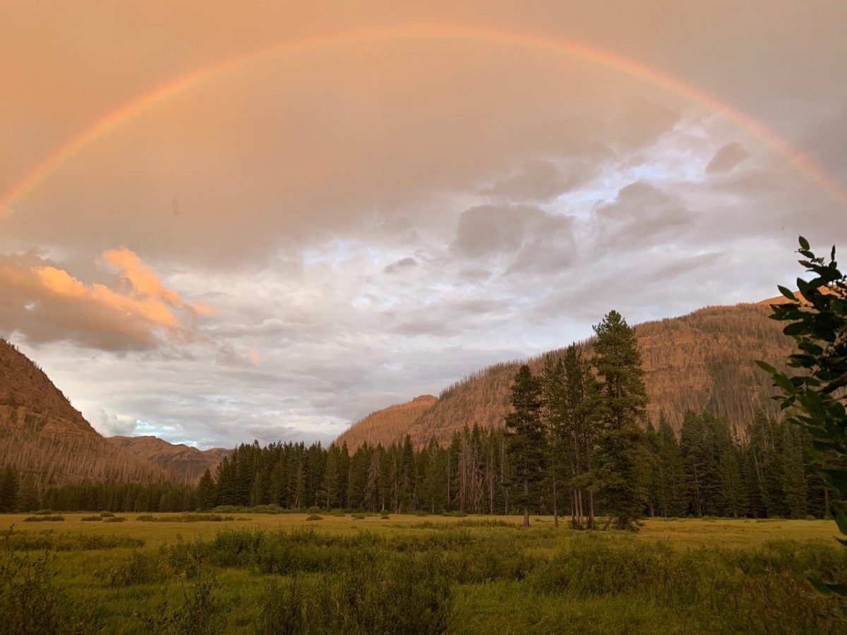 A landscape view of mountains in Wyoming, with a field in the foreground and a rainbow in the sky