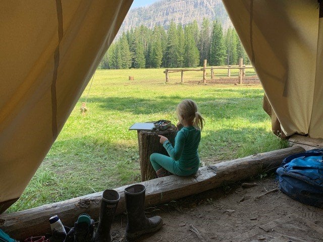 A girl sitting at the entrance to a canvas tent sitting on a log