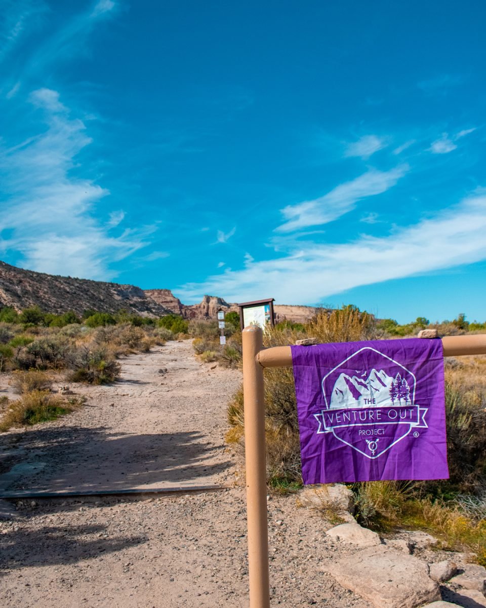 A purple Venture Out Project flag hanging on a wooden post, with a desert trail in the background