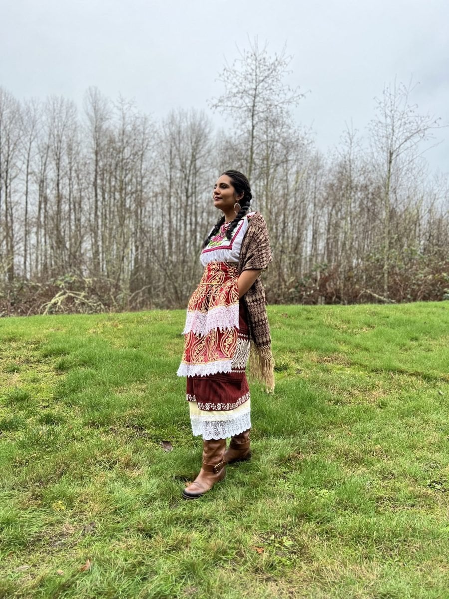 A woman dressed in traditional P'urhepecha regalia standing in a grassy field, with bare trees behind her