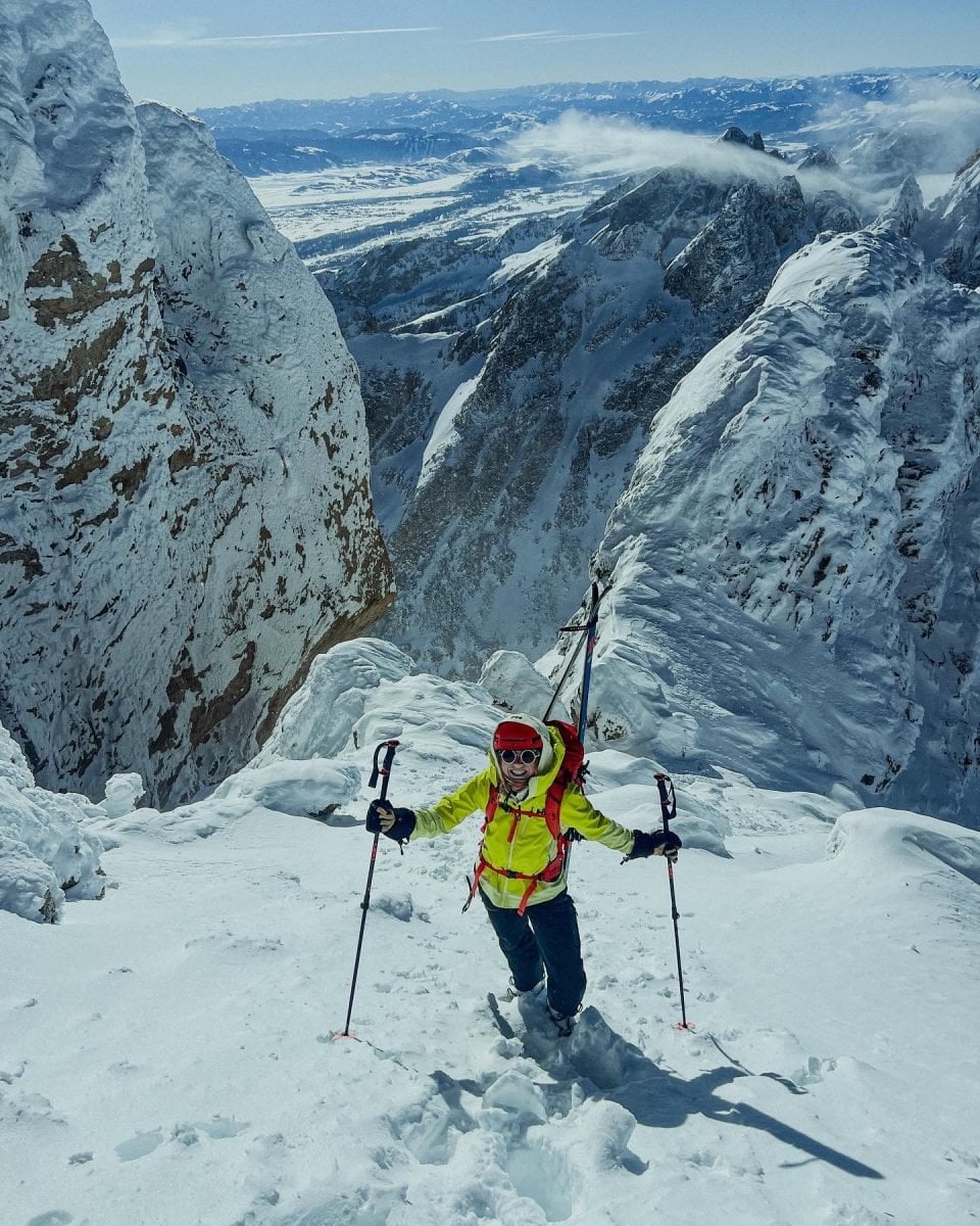A skier in yellow and orange gear posing in gear atop a snowy mountain