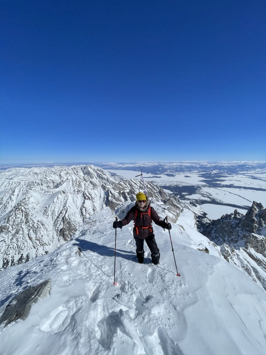 A woman in ski gear standing on a snowy mountain peak, with snowy landscape in the distance