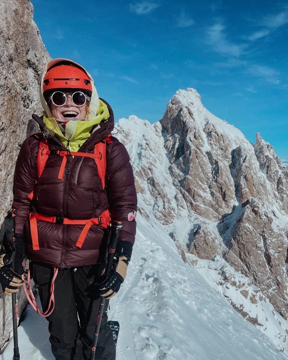 A woman in full ski gear standing among mountain peaks