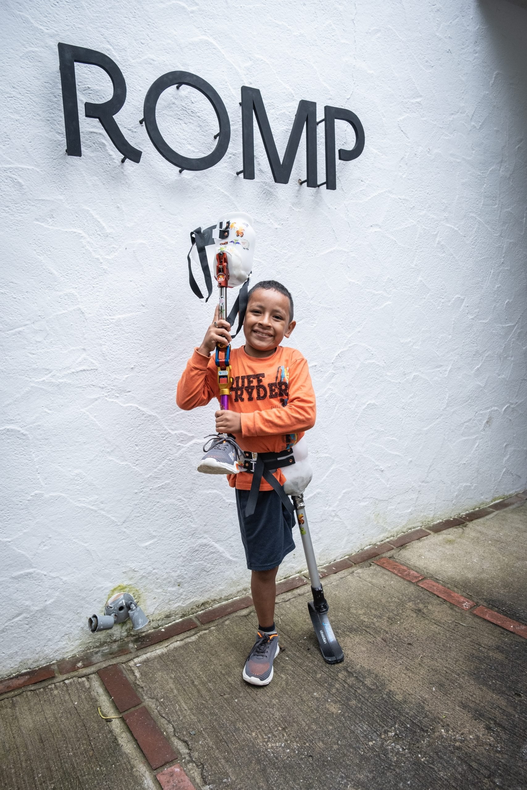 A child wearing an orange shirt holding a prosthetic running leg while standing under a ROMP sign