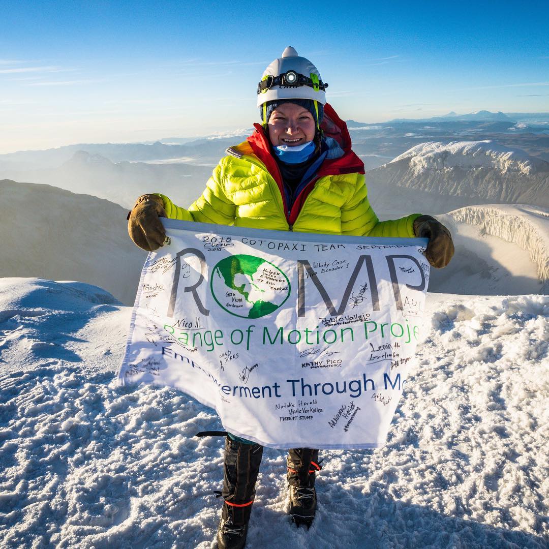 A woman standing in cold-weather gear atop a snow-covered mountain holding a ROMP flag with names and signatures covering it