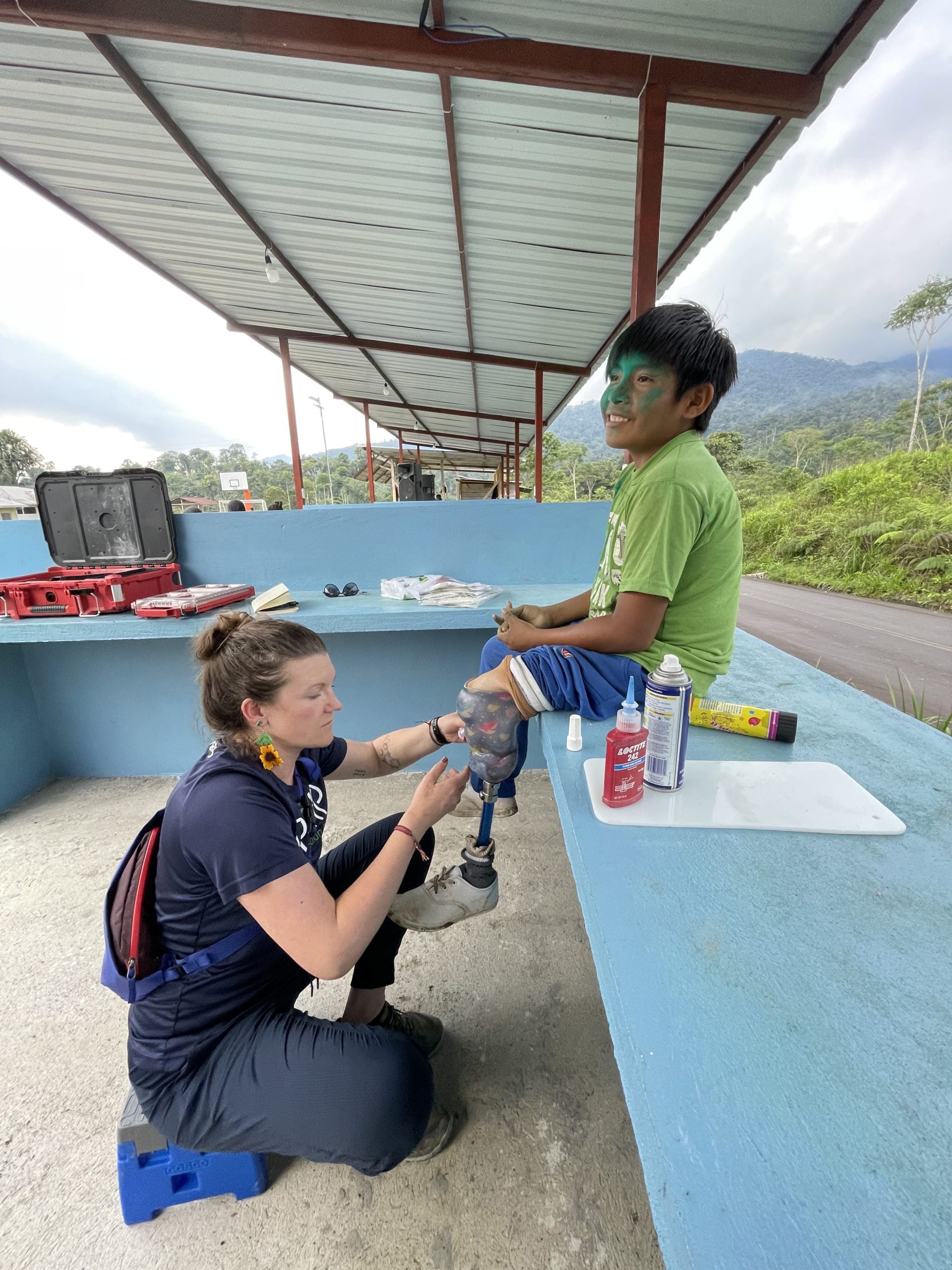 A woman sitting on a stool on the ground fitting a young boy with a prosthetic
