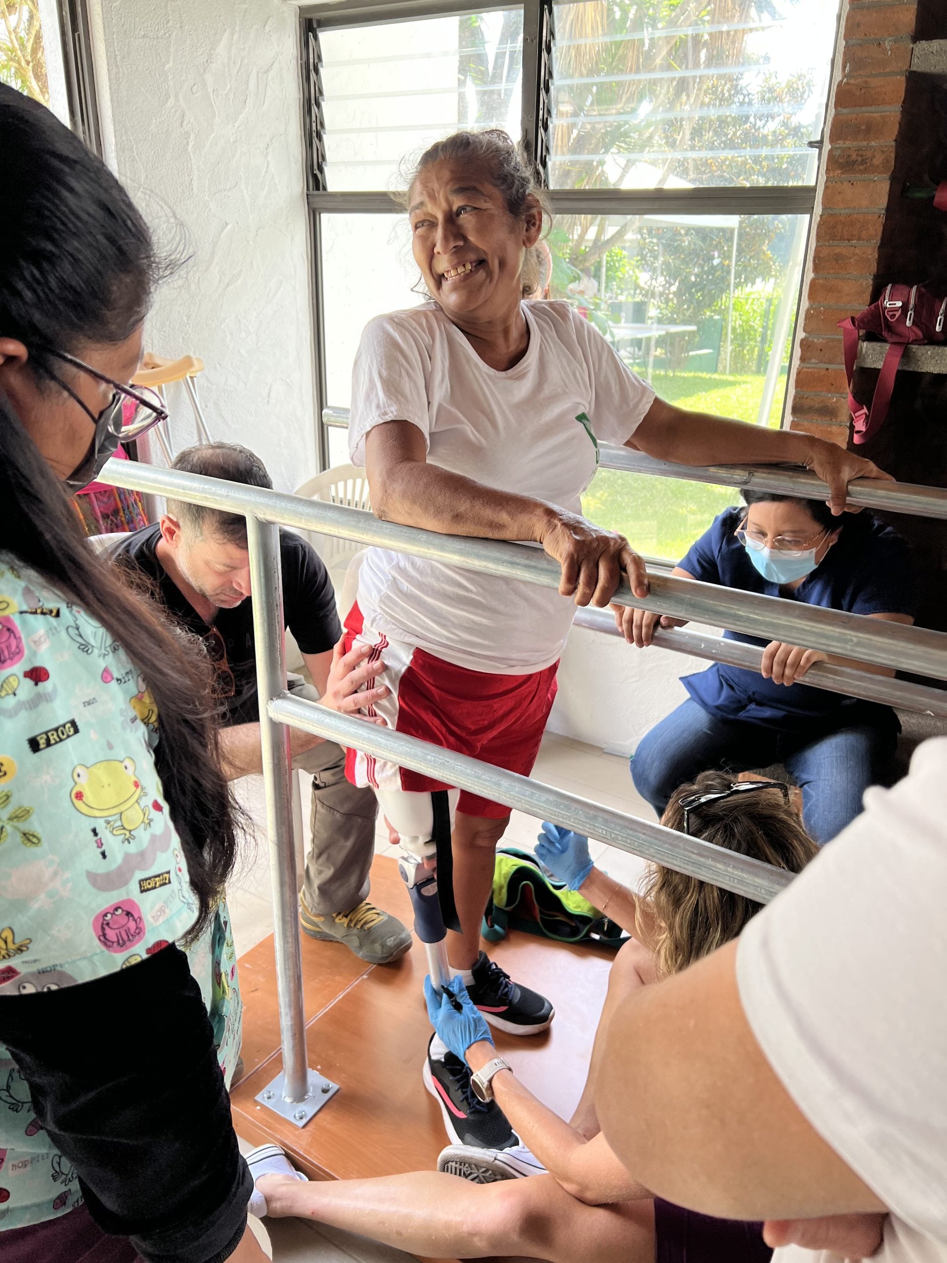 A woman smiling and standing between two sets of metal bars with a prosthetic leg, with folks standing around supporting her.