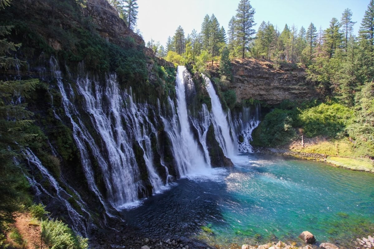 A large cliff face with waterfall(s) falling across it, and a blue swimming hole below