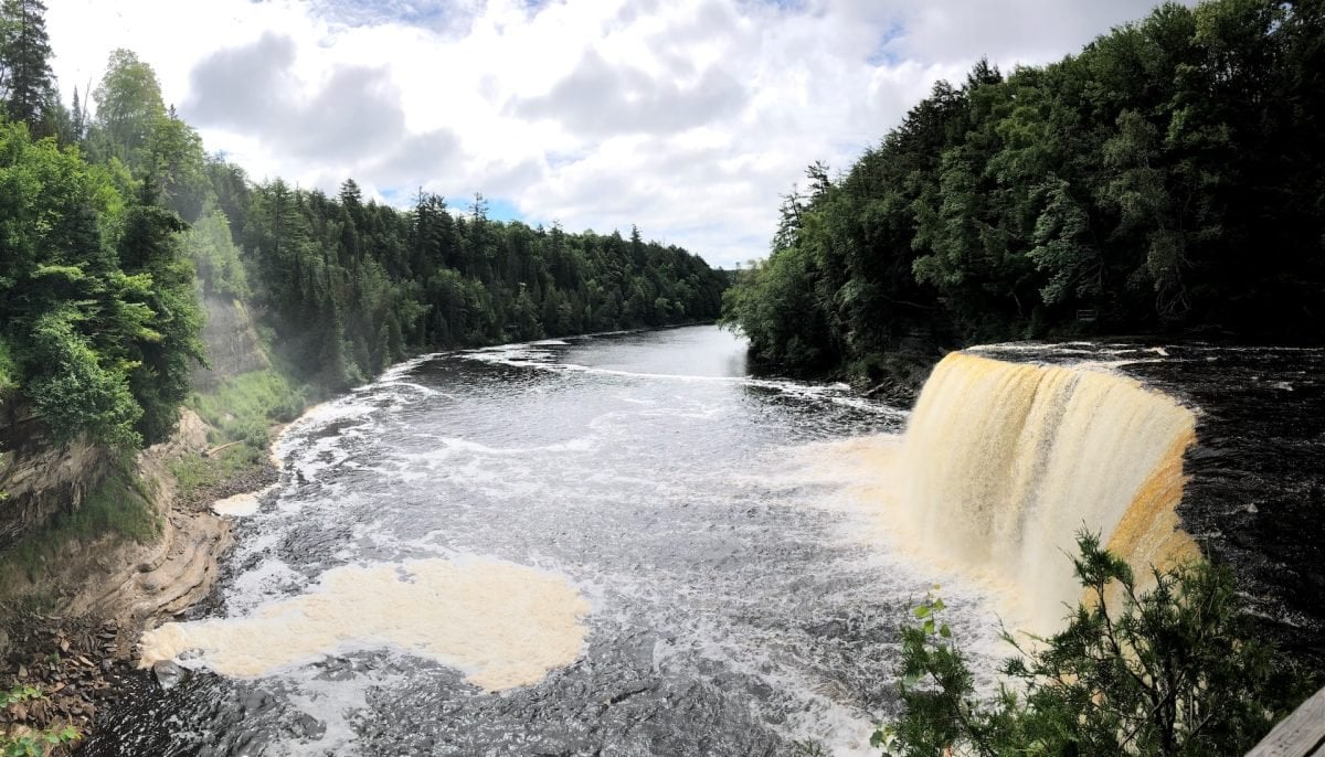 A waterfall with a light brown hue falling into a body of water below, and surrounded by forested banks on either side