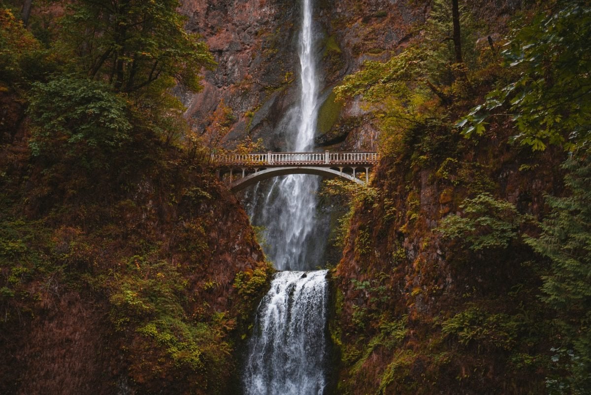 A two-tiered waterfall with a bridge overlooking both, surrounded by moss and trees on either side