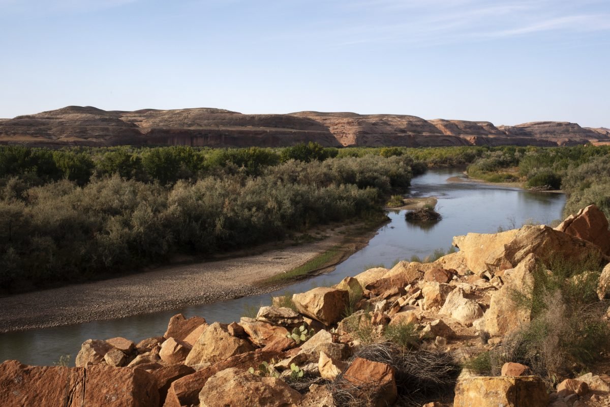 A sweeping view of desert landscape, with rocks and a river in the foreground and rolling mountains in the background