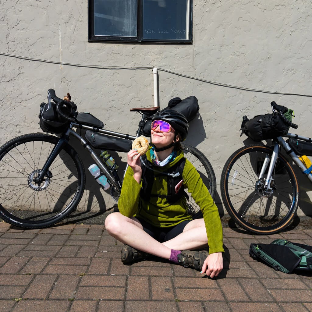 A cyclist sits on the ground in the sunlight holding a treasured bagel with bikes leaning against a building in the background.