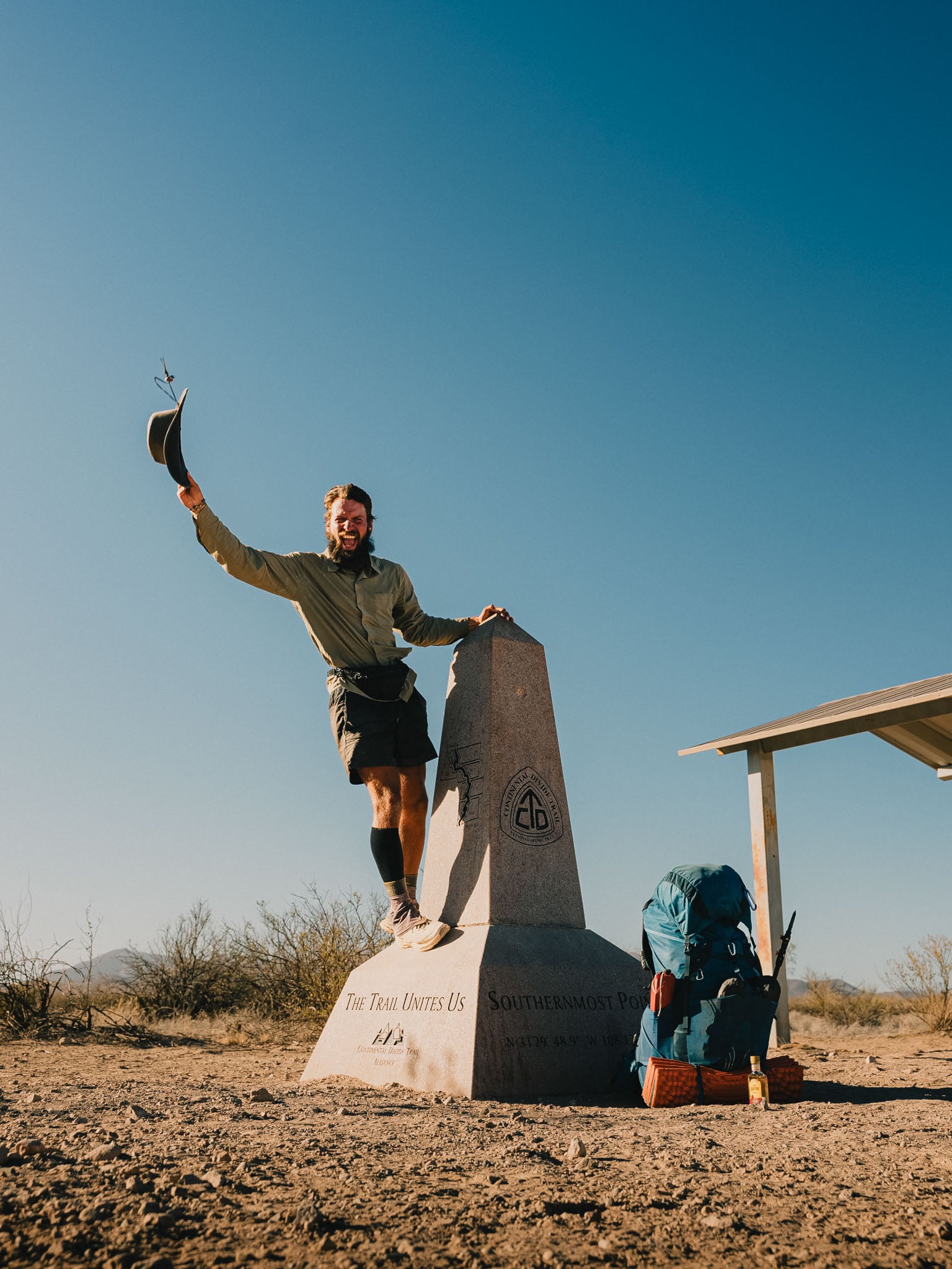 A hiker poses with a CDT marker, waving his hat in the air.