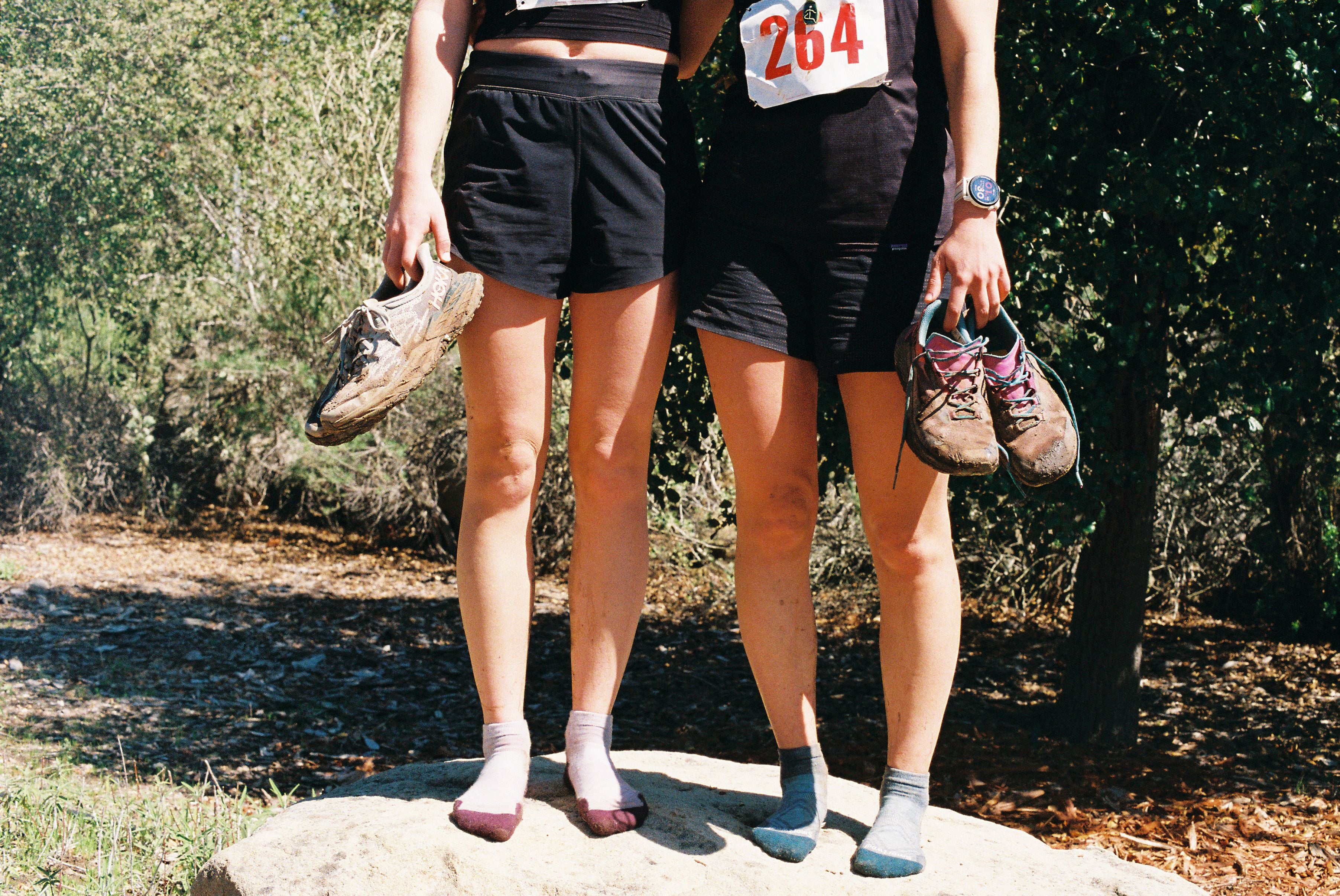 Two runners stand side by side with their muddy shoes in hand.