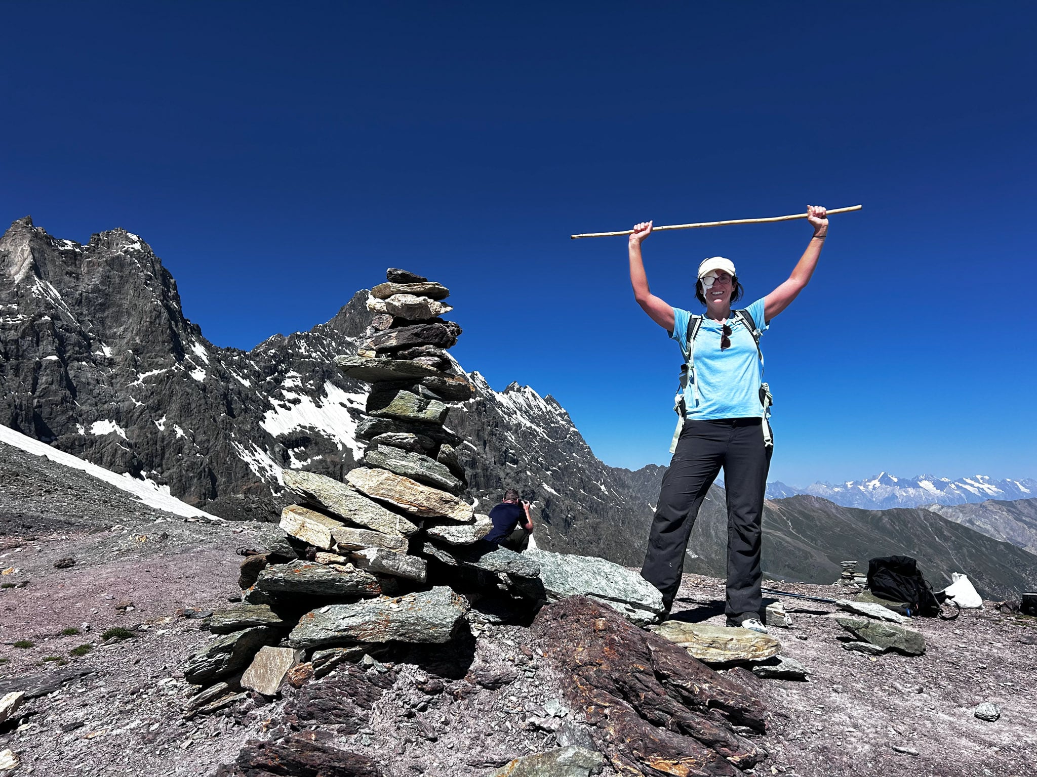 A hiker stands next to a rock cairn raising their walking stick triumphantly above their head.