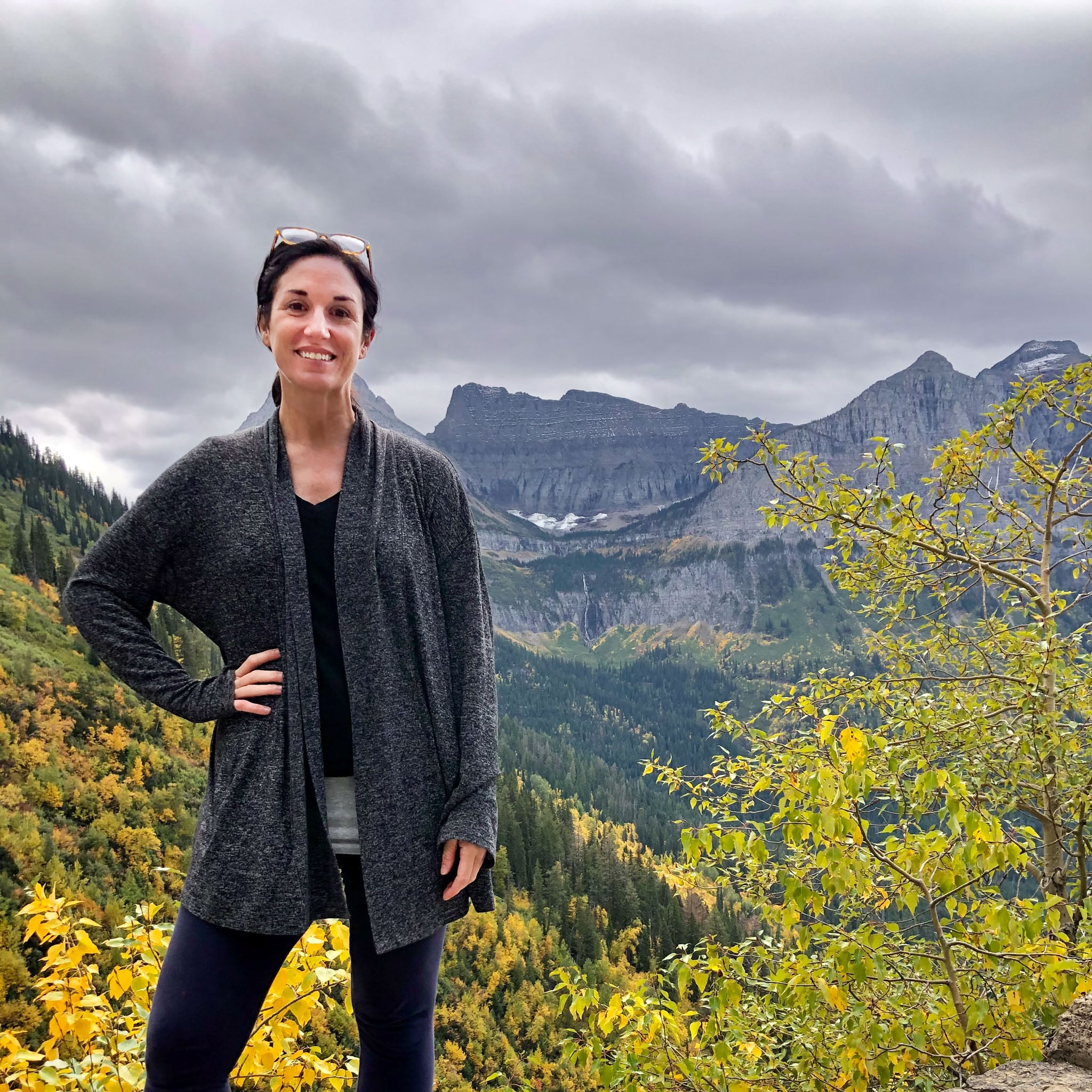 Author, Laura Kiniry, poses in front of a vista of mountains and trees in Glacier National Park.