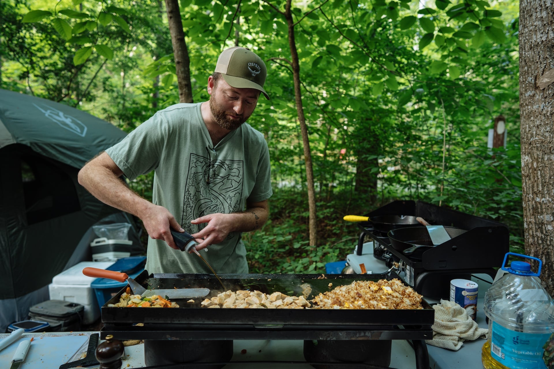 A person cooks over a griddle in the forest.