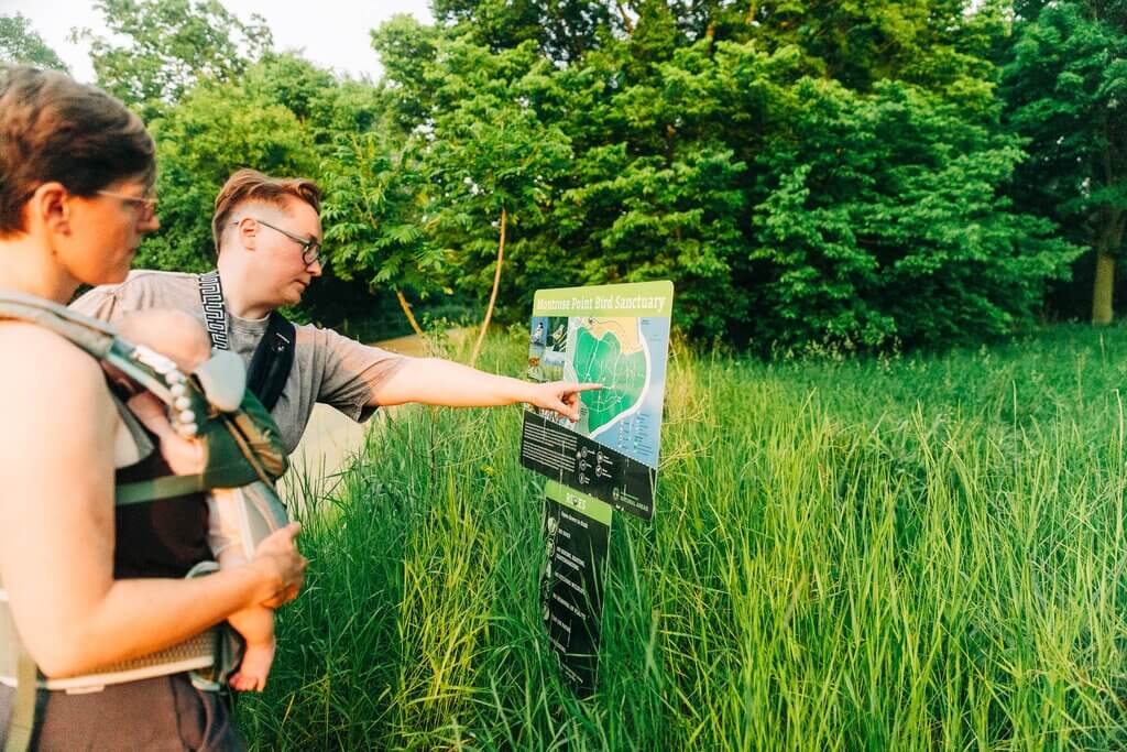 A couple stands at a park map. One person has an infant held in a front carry baby carrier and the other points to a route on the map.