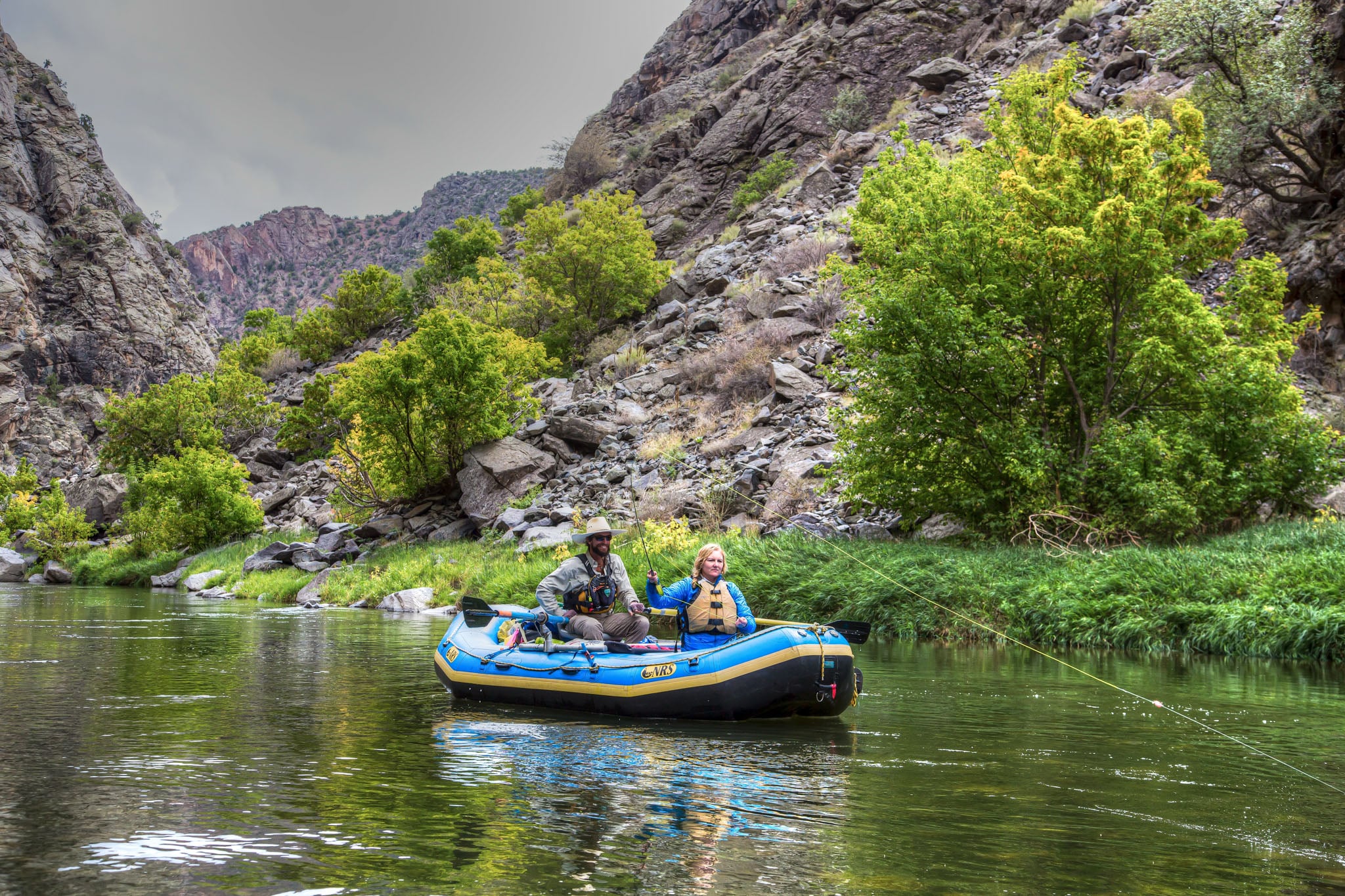 Two people fish from an inflatable raft on a river lined with trees.