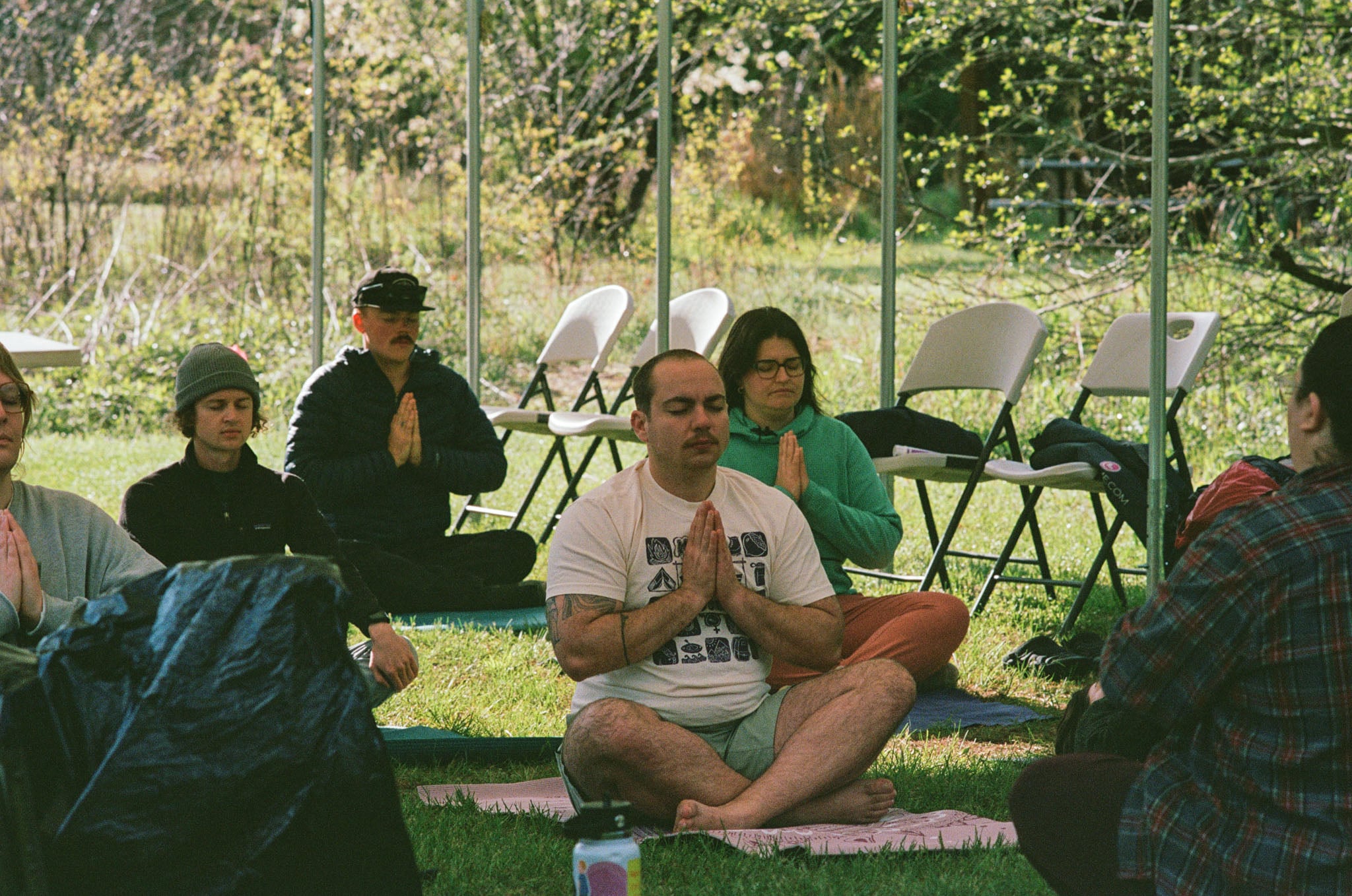 A group of people sit for yoga practice under a tent in the grass.