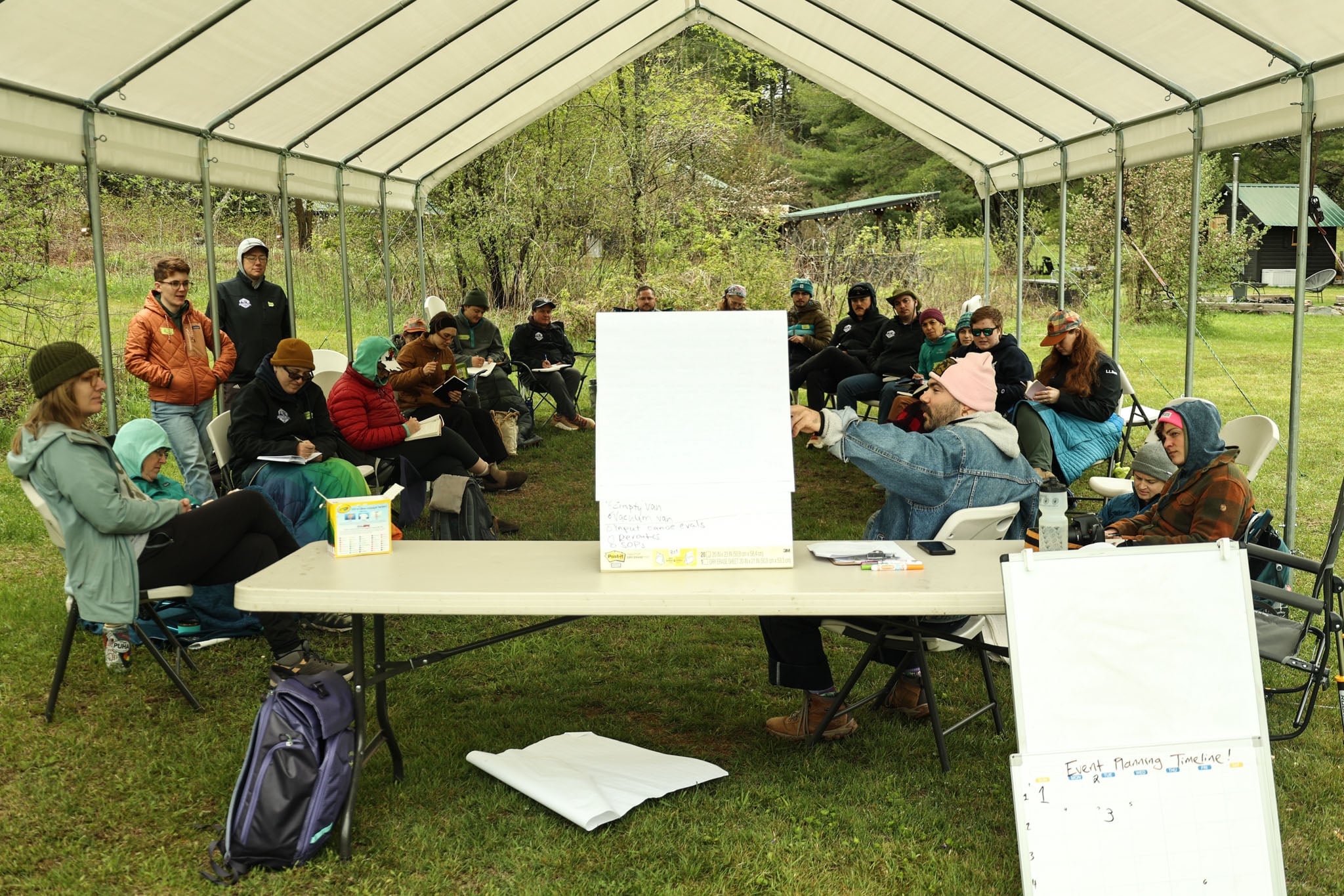 A group of people gather under a tent and take notes during a presentation.