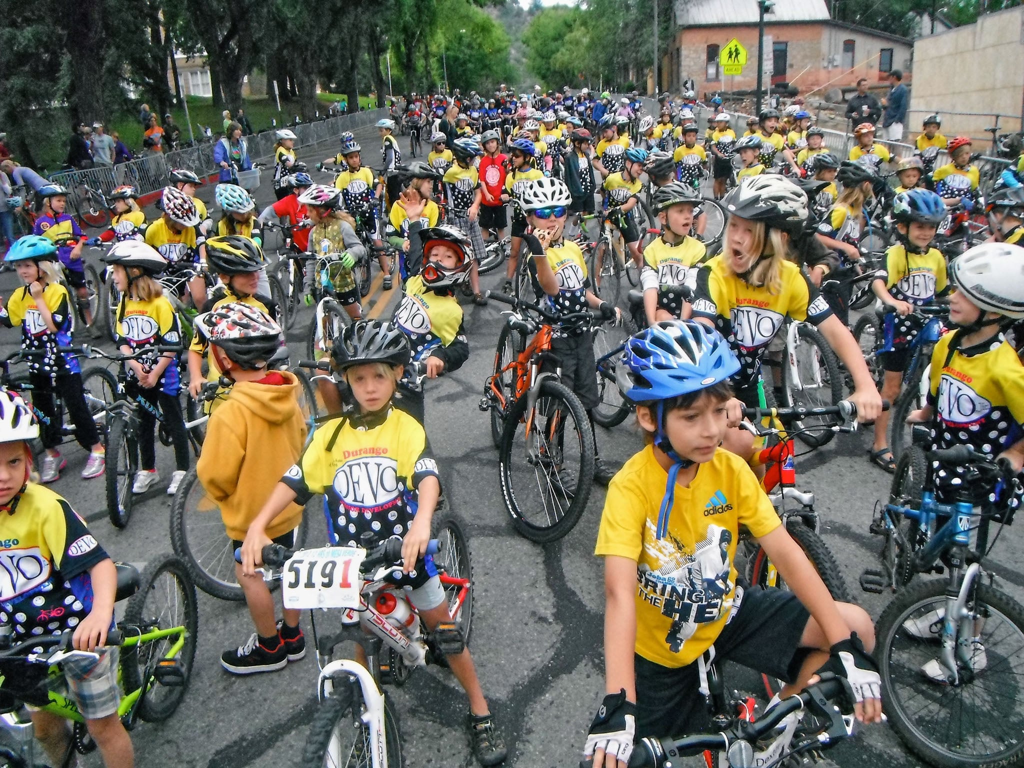 A group of kids on a street in helmets and yellow Devo shirts on bicycles.