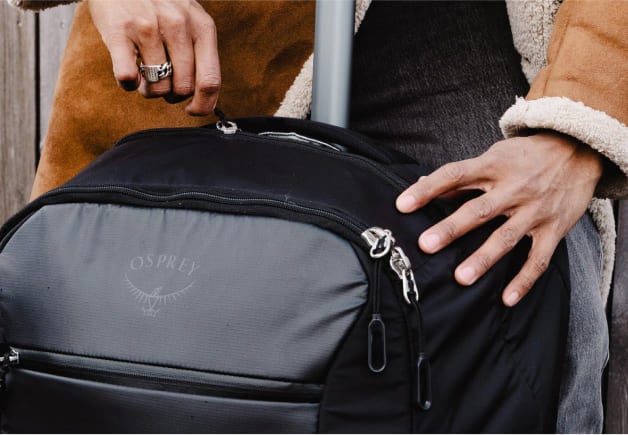 Close-up of a person&rsquo;s hands holding the zippers of a black Osprey travel bag with a visible brand logo.