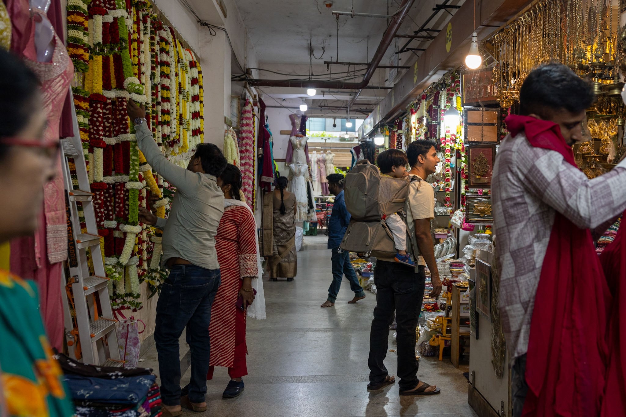 A person walks through a busy marketplace with a child on their back in a child carrier.