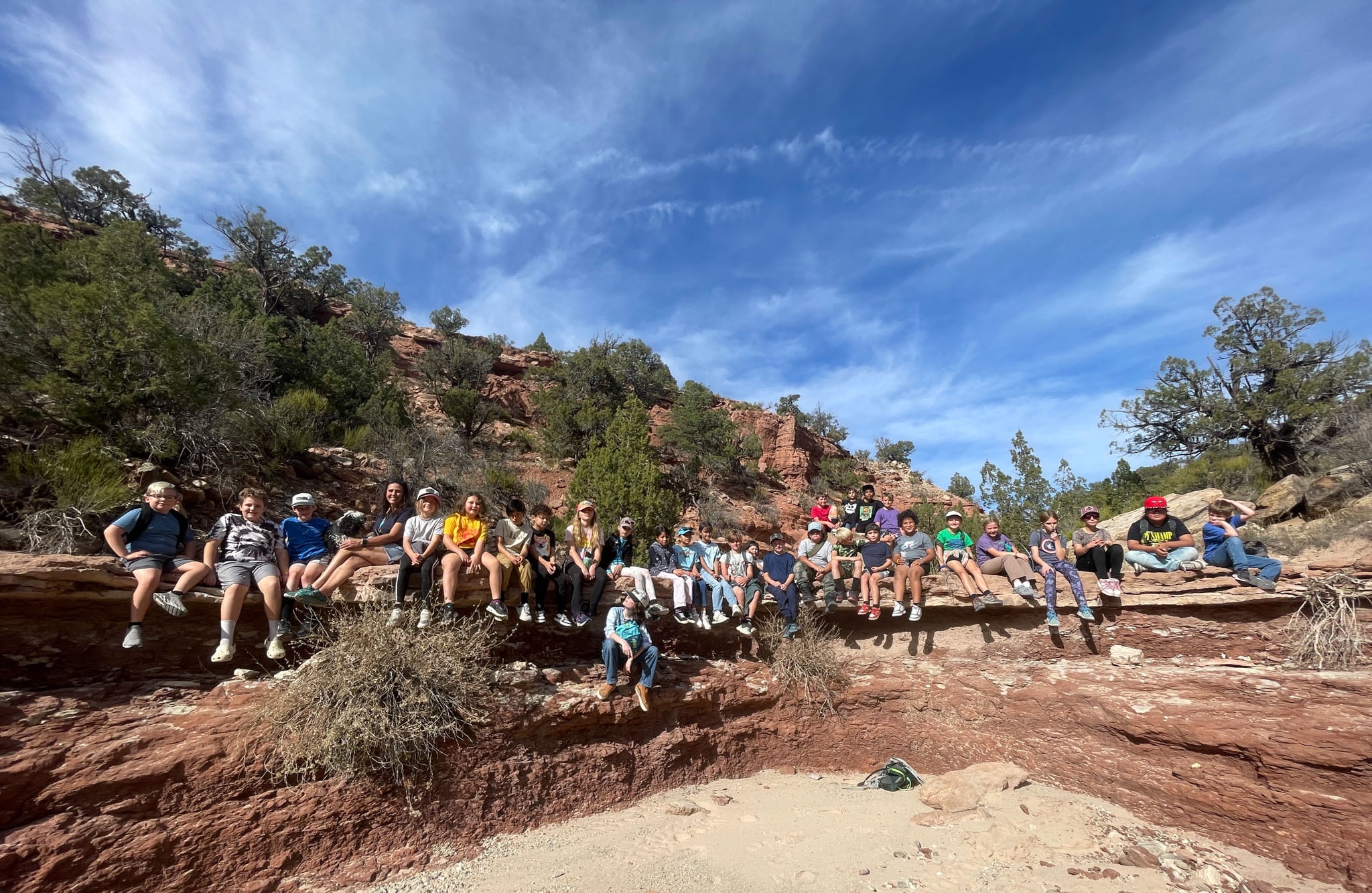 A group of students sit on a rock overhang in a desert wash.