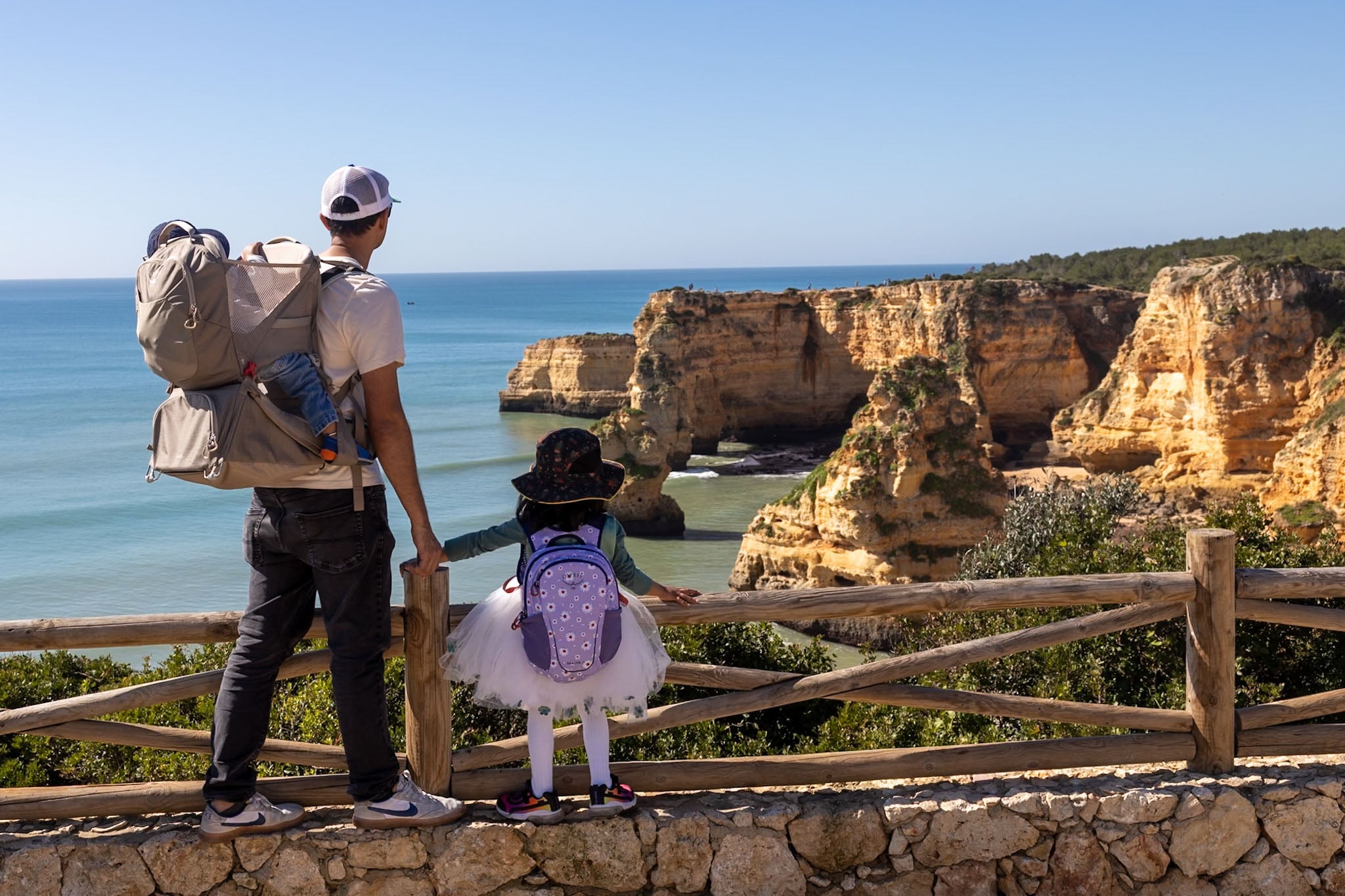 An adult with a child on their back in a child carrier and a child on foot holding their hand look over a rocky sea scape.