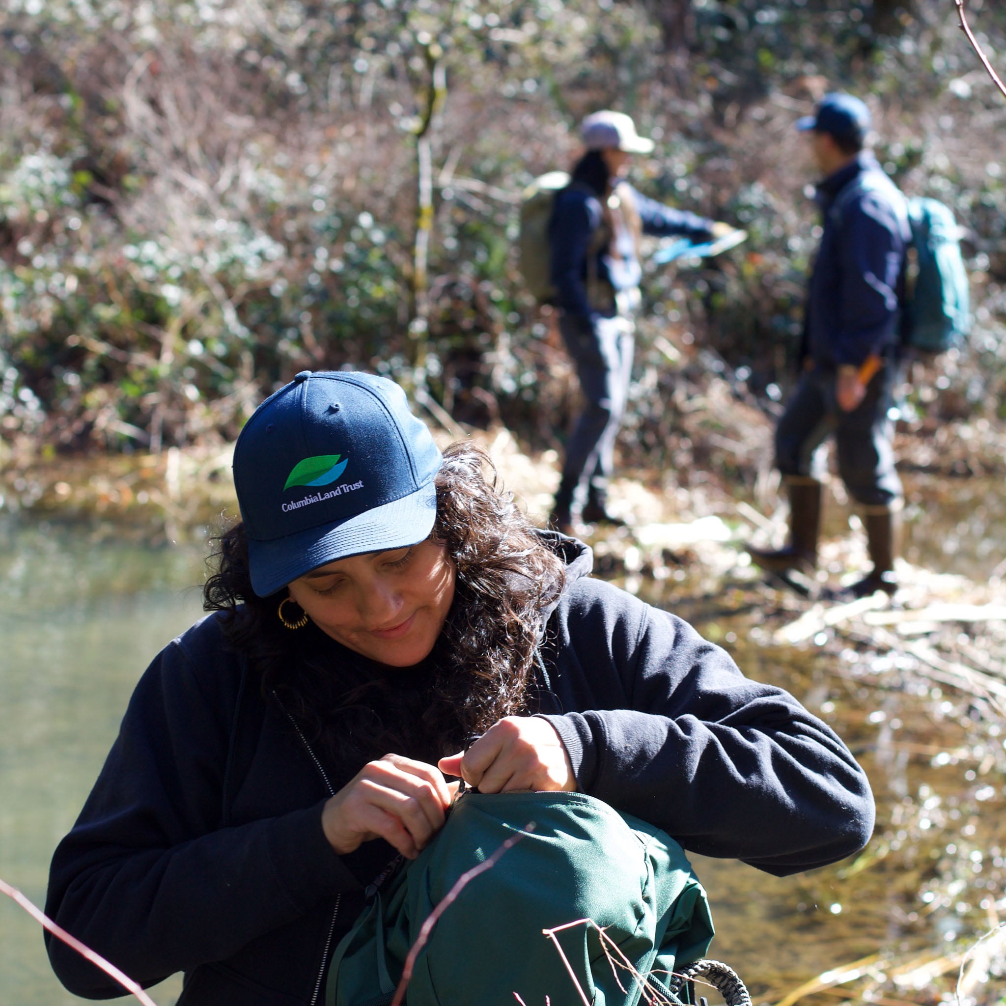 A person closes their backpack, two people talk in the background.