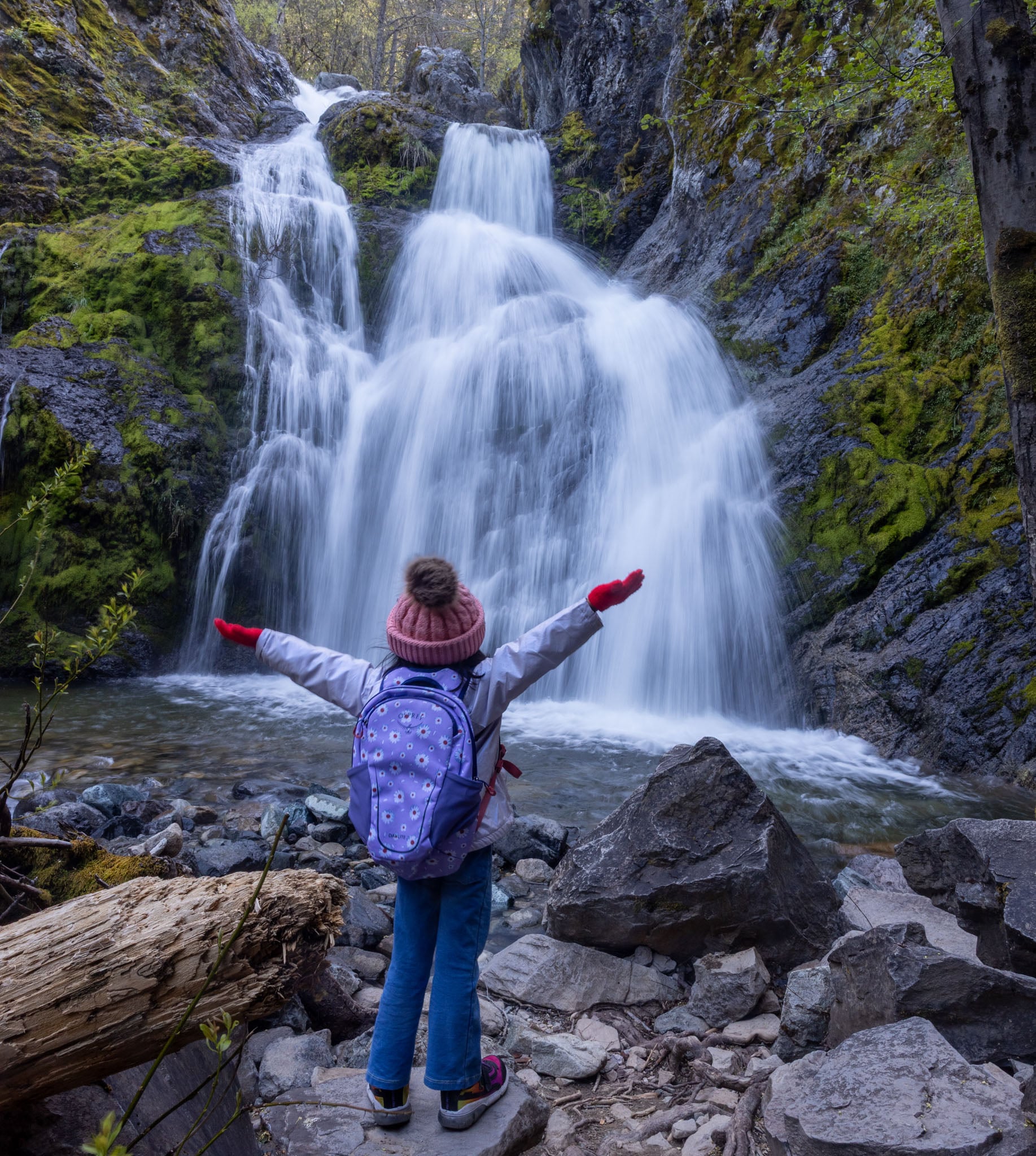 A child stands in front of a waterfall, arms outstretched.