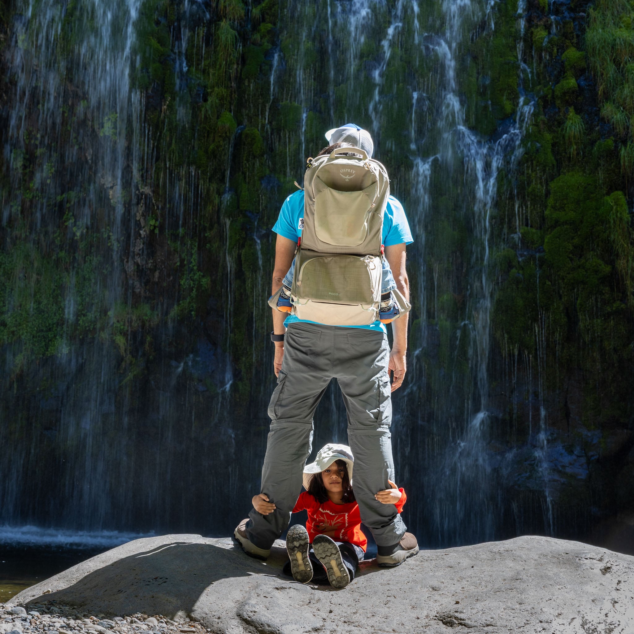A person and two children stand in front of a waterfall, one child riding in a child carrier on the adult's back, one child sits between the adult's legs looking at the camera.