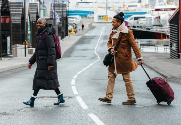 Two people walking across a street near a harbor, one carrying a maroon backpack and the other pulling a matching maroon wheeled suitcase.